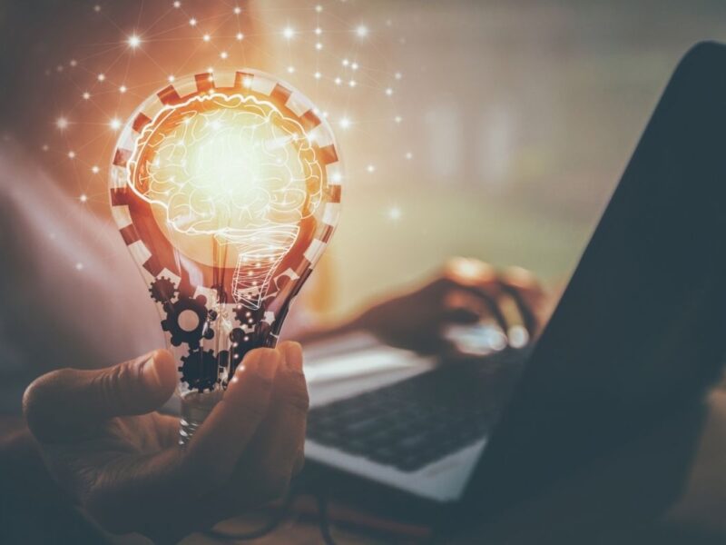 Mann arbeitet an einem Laptop und hält in einer Hand eine leuchtende Glühbirne, in der Glühbirne zeichnet sich ein Gehirn ab
